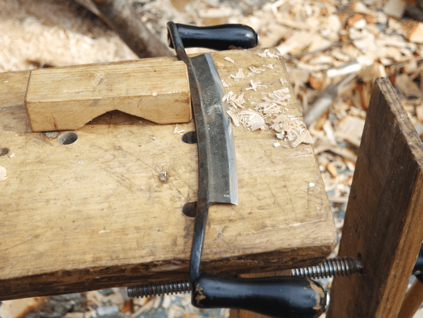 curved drawknife with black handles lying on a piece of timber with timber shaving scattered around.